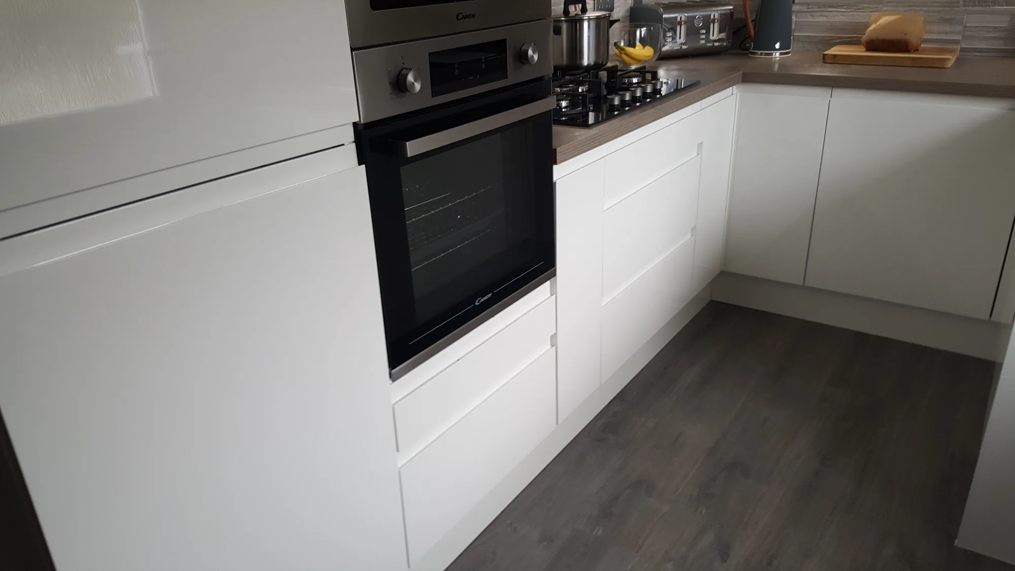 J-pull drawer stacks and white gloss doors below the hob run, with grey woodgrain flooring