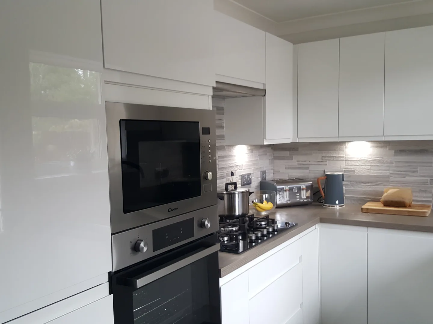 White gloss handleless kitchen in Glasgow with a raised oven and microwave, gas hob and stone-effect splashback