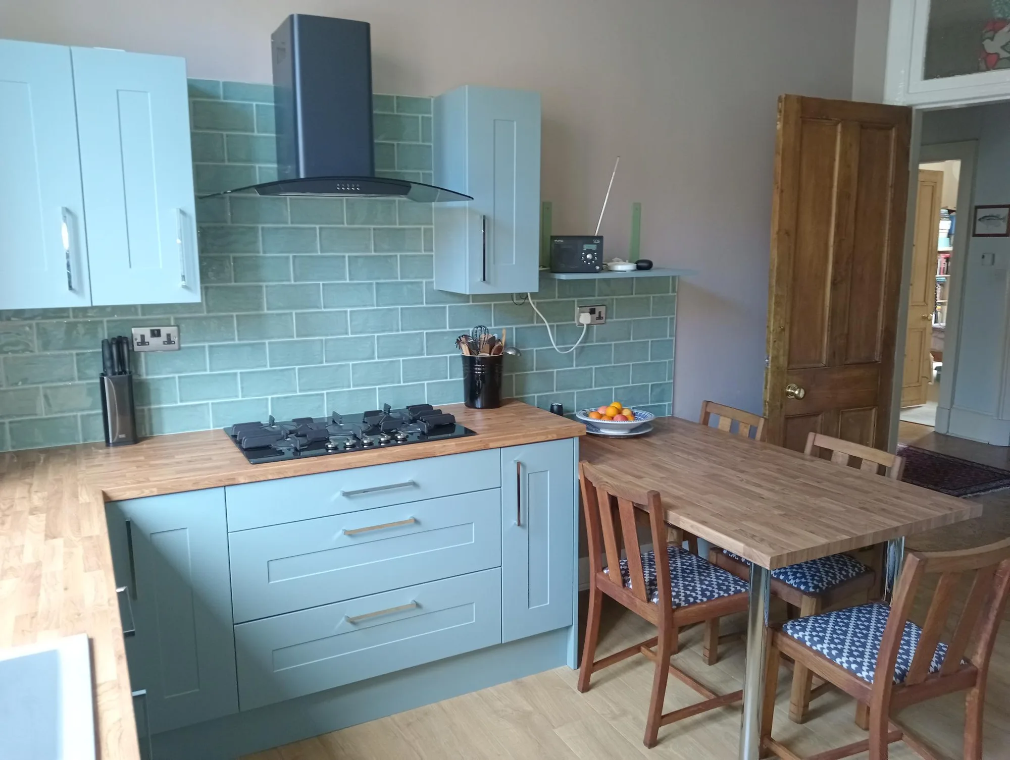 Gas hob under a curved glass chimney hood with green metro tiles and the family table alongside, in a Glasgow tenement kitchen