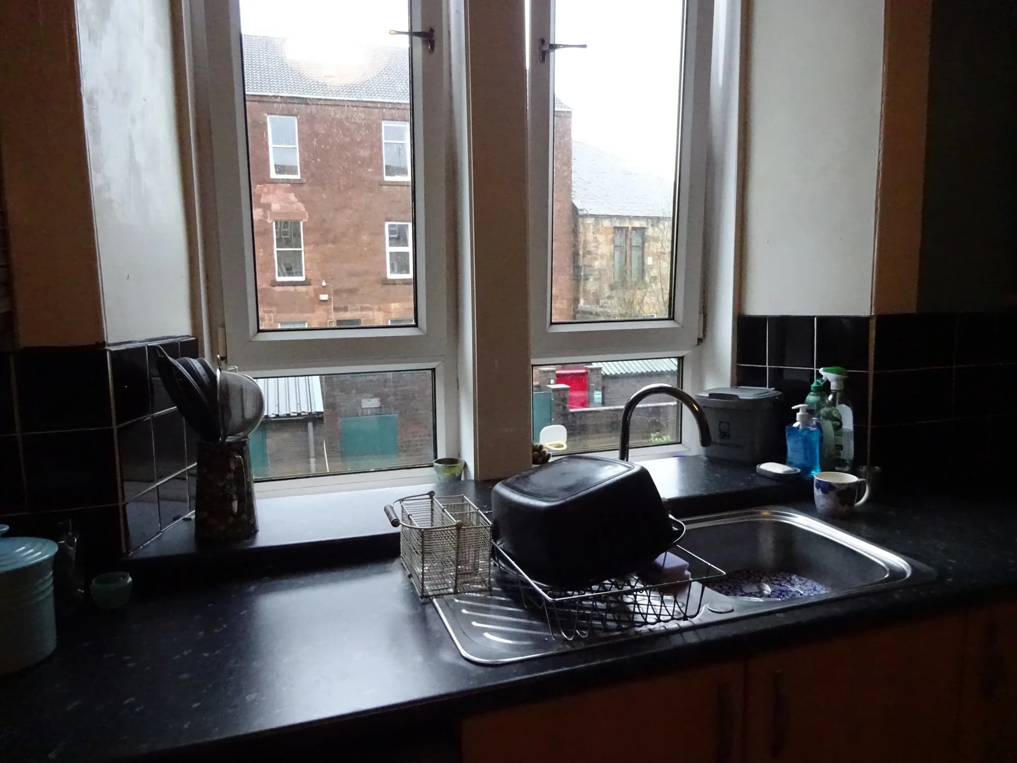 The old sink under the tenement window before renovation, with black tiles wrapping the reveal and a dark laminate worktop