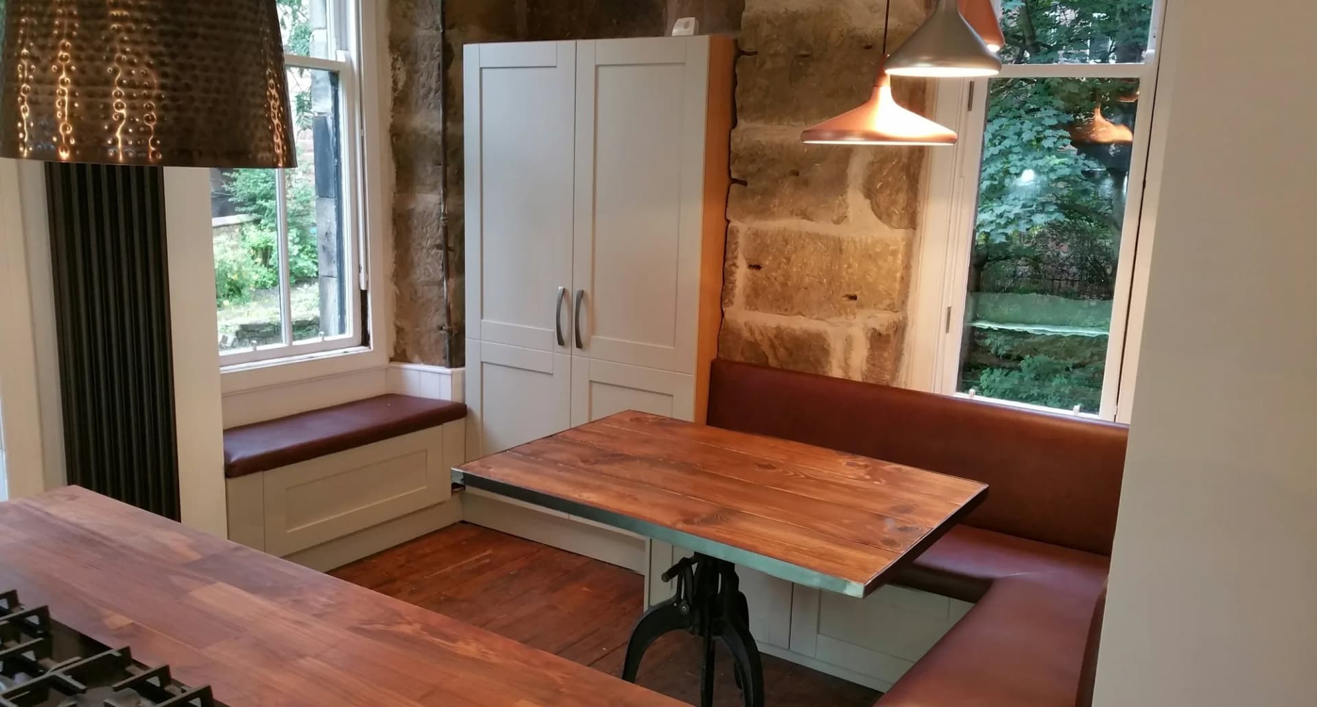 Leather banquette and window seat under two sash windows beside an exposed sandstone wall, with a tall shaker cupboard and copper pendants
