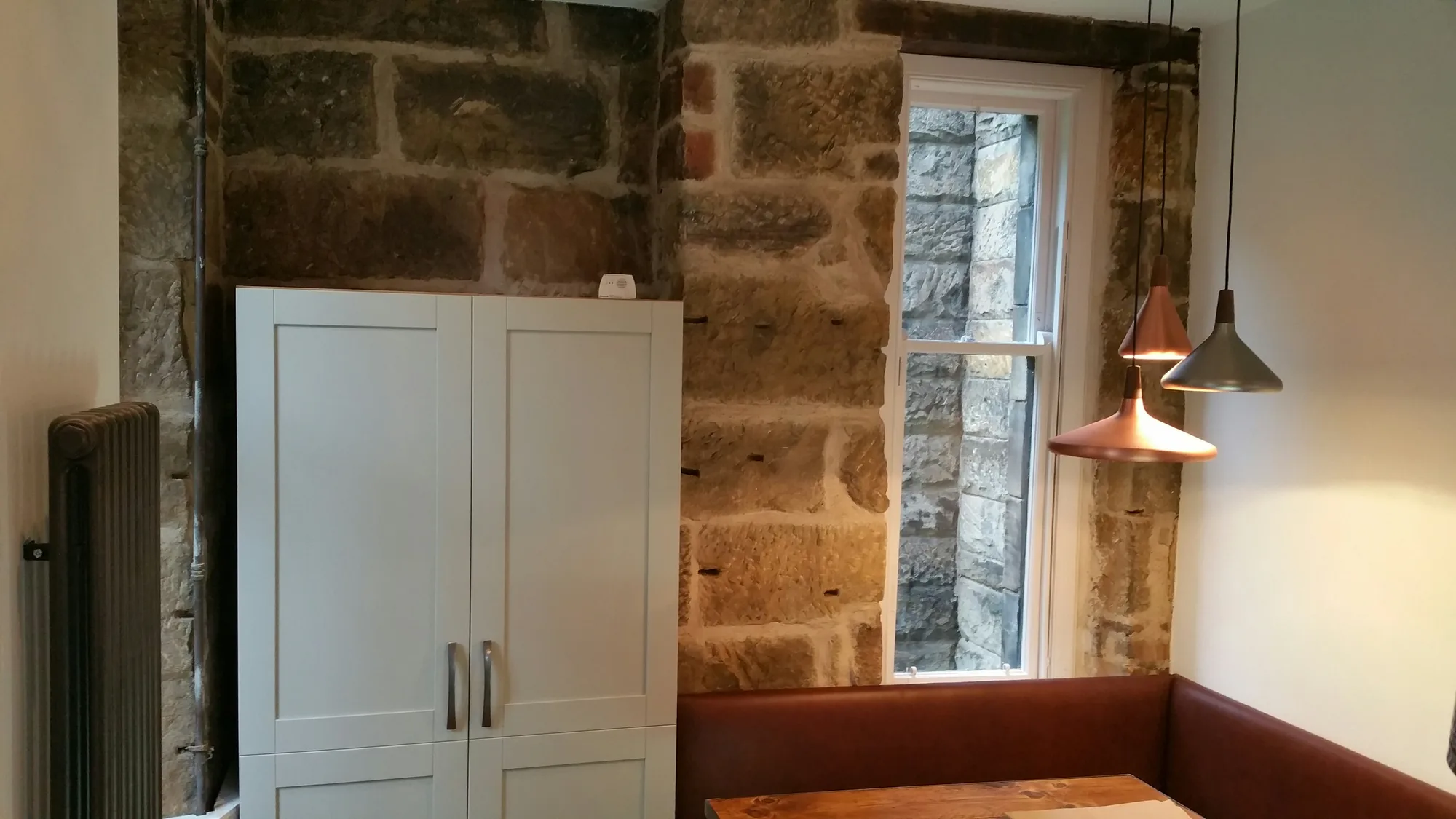 Tall off-white shaker cupboard against a bare sandstone wall, beside a sash window and a cluster of copper pendant lights