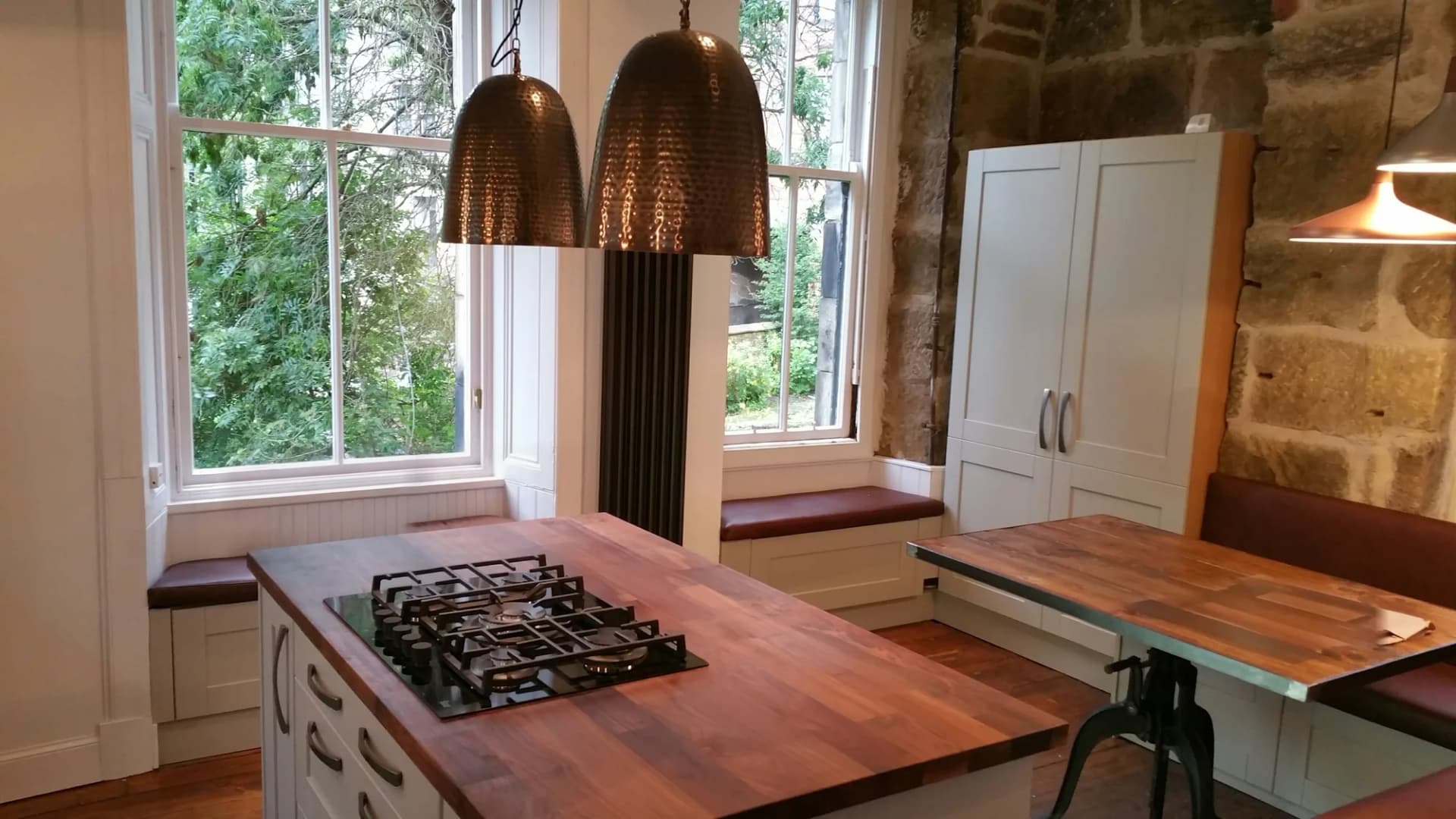 Timber-topped kitchen island with a gas hob under hammered bronze pendants, looking towards the sash windows and the window seat