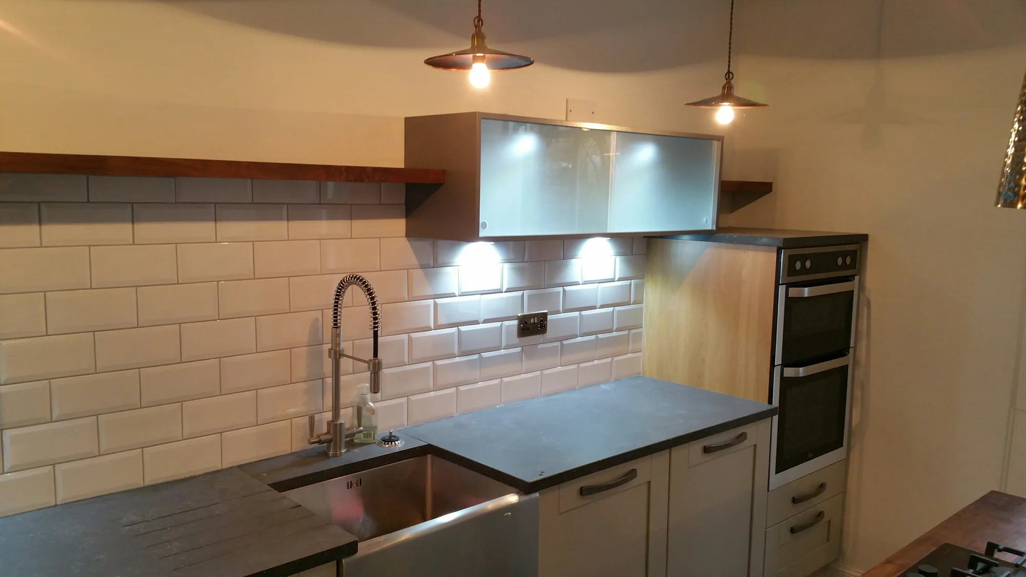 Stainless steel apron-front sink with a pull-down spring tap and drainer grooves in a dark worktop, with white metro tiles and a stainless wall unit above