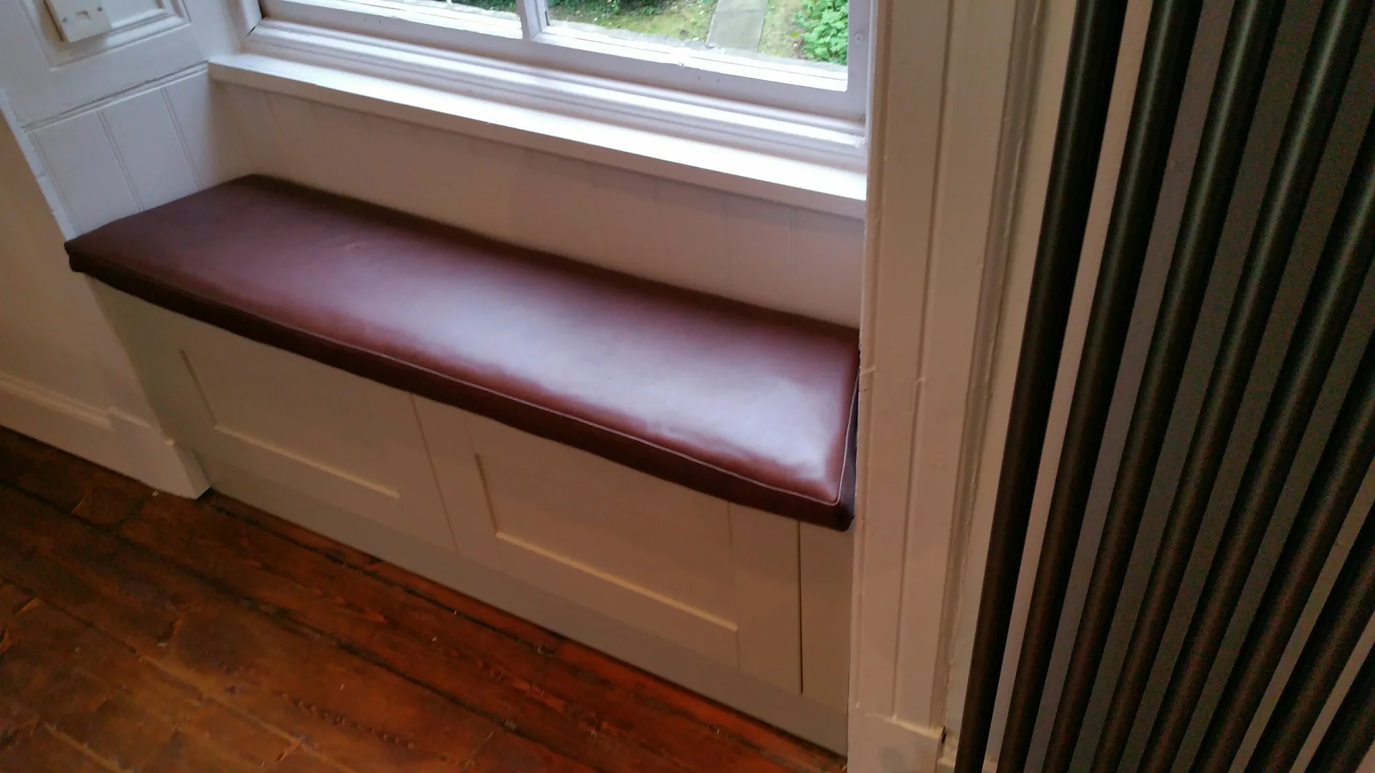 Built-in window seat with an oxblood leather cushion on a panelled shaker base, beside a tall column radiator on stripped floorboards